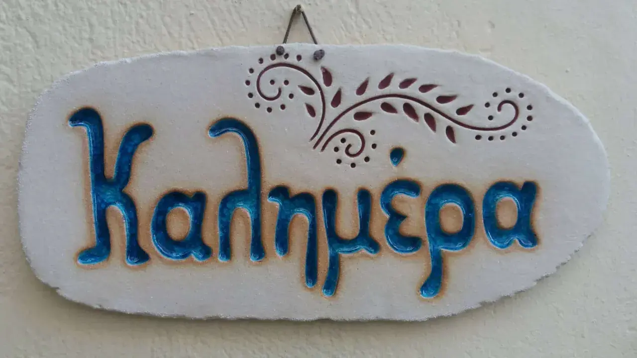 Ceramiczna tabliczka z napisem "Καλημέρα" (do zobaczenia po grecku) i ozdobnym motywem roślinnym.