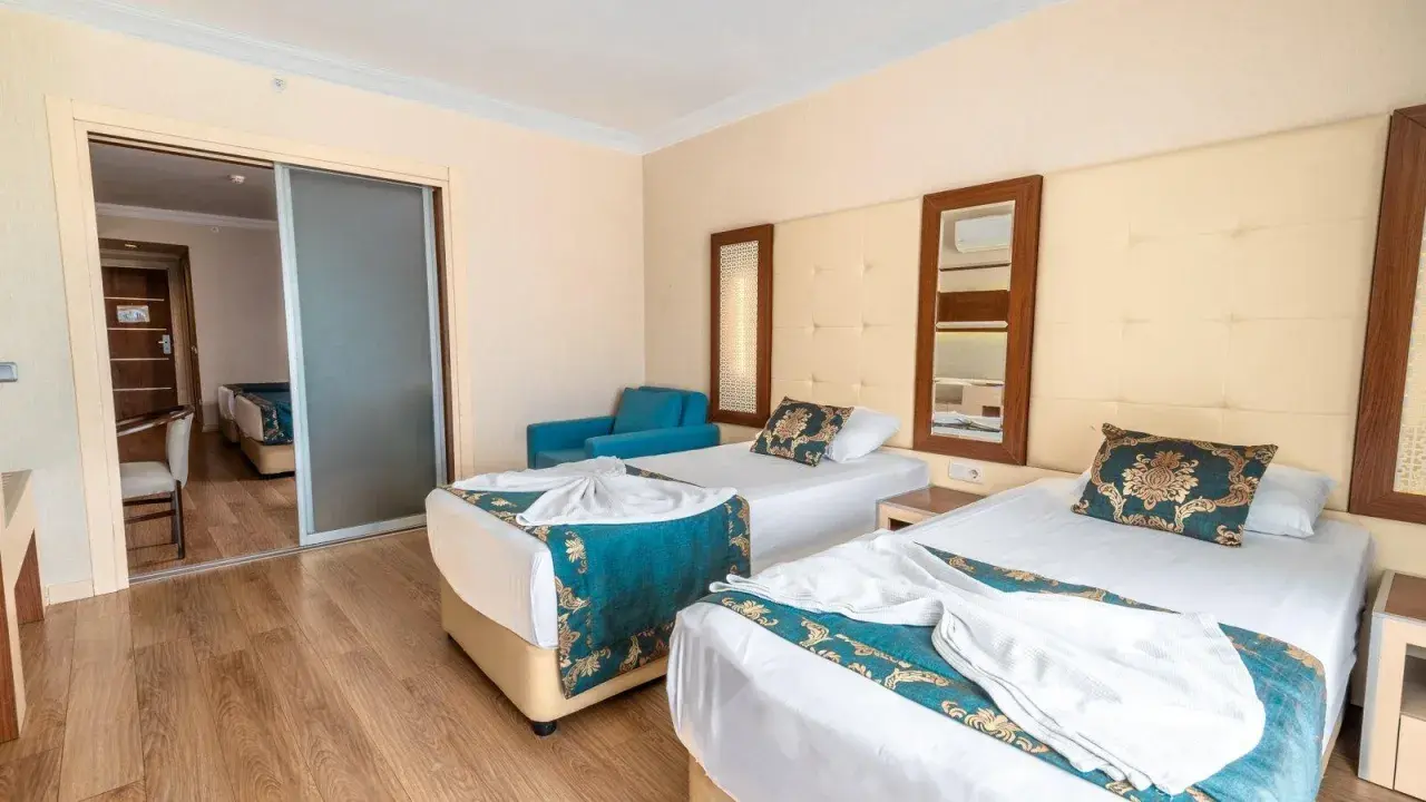 Pokój hotelowy z dwoma łóżkami, ozdobiony turkusowymi narzutami. Widok na sąsiedni pokój przez przesuwne drzwi. Idealne miejsce na relaks po dniu spędzonym w beach club doganay.