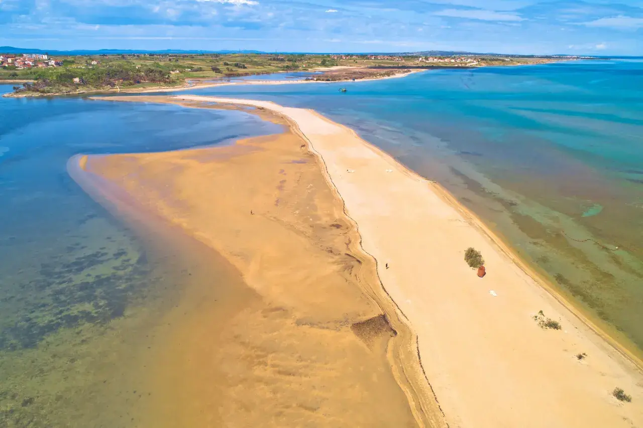 Chorwacja plaże piaszczyste, piaszczysta mierzeja otoczona turkusową wodą, w oddali widać zabudowania.