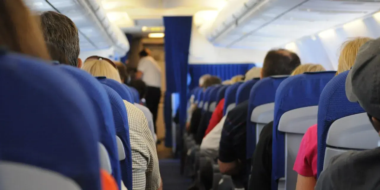 Widok z perspektywy pasażera w samolocie. Siedzący ludzie, niebieskie siedzenia, stewardessa w tle. To może być inspiracja, jak nie bać się latać.