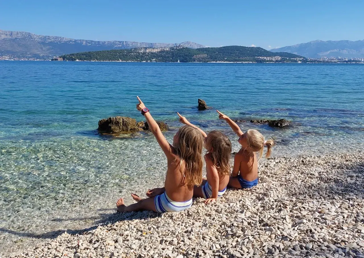 Trzy dziewczynki siedzą na kamienistej plaży, wskazując palcami na morze i góry. Mówią sobie "Dzień dobry po chorwacku" w słoneczny dzień.