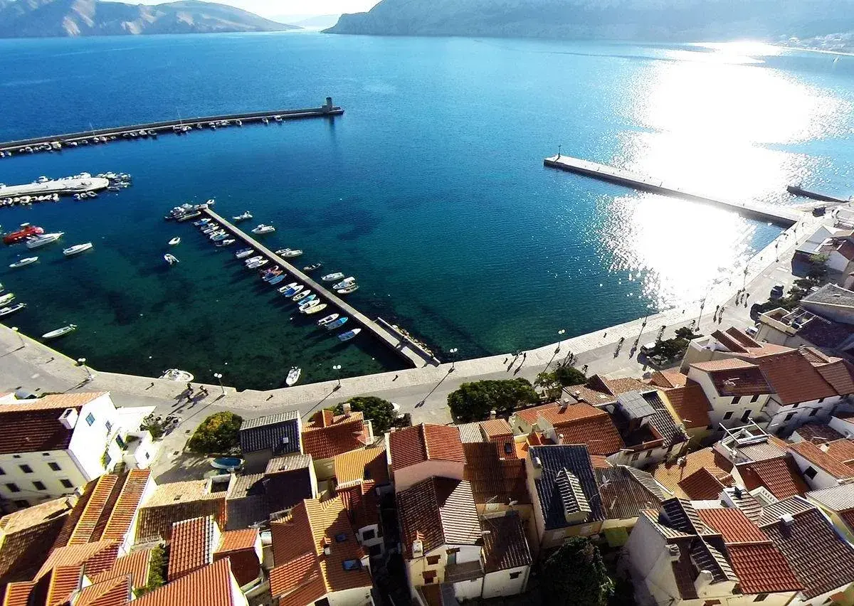 Widok na port i miasteczko Baška w Chorwacji. Woda jest turkusowa, a na nabrzeżu cumują łodzie.