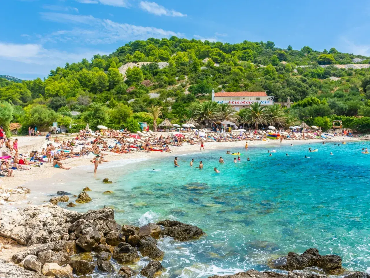 Słoneczna plaża w Chorwacji, ludzie opalają się i kąpią w krystalicznie czystej wodzie. Idealne miejsce na wakacje all inclusive samolotem.