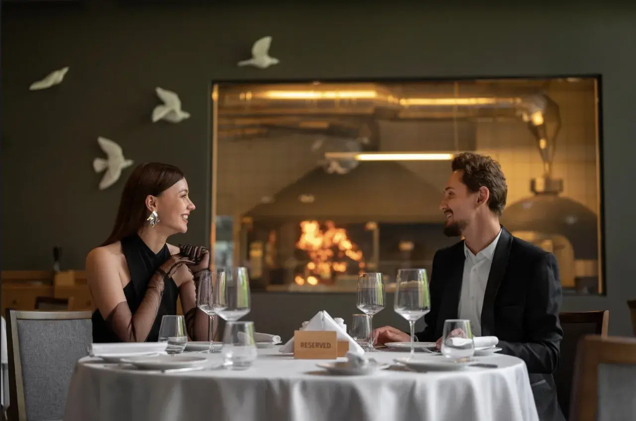 Elegancka para cieszy się romantyczną kolacją w restauracji a la carte all inclusive. Za nimi widać kuchnię z ogniem w piecu.