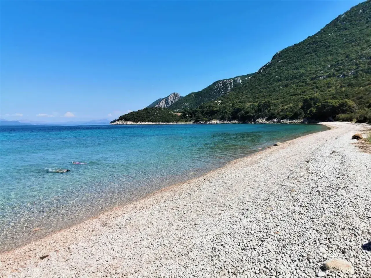 Chorwacja piaszczyste plaże blisko Polski. Dwie osoby pływają w krystalicznie czystej wodzie przy kamienistej plaży, w tle zielone wzgórza.