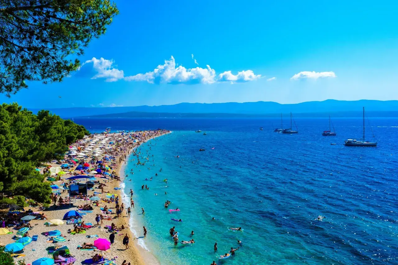 Wyspa Brać oferuje niezapomniane atrakcje: plaża Zlatni Rat tętni życiem, ludzie kąpią się w turkusowej wodzie, a żaglówki kołyszą się na morzu.