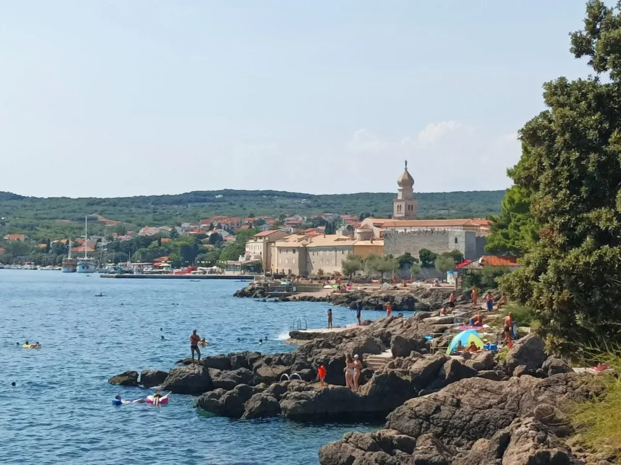 Słoneczny dzień na wyspie Krk. Ludzie odpoczywają na skalistej plaży, kąpią się w morzu. W tle widać miasteczko z charakterystyczną wieżą.
