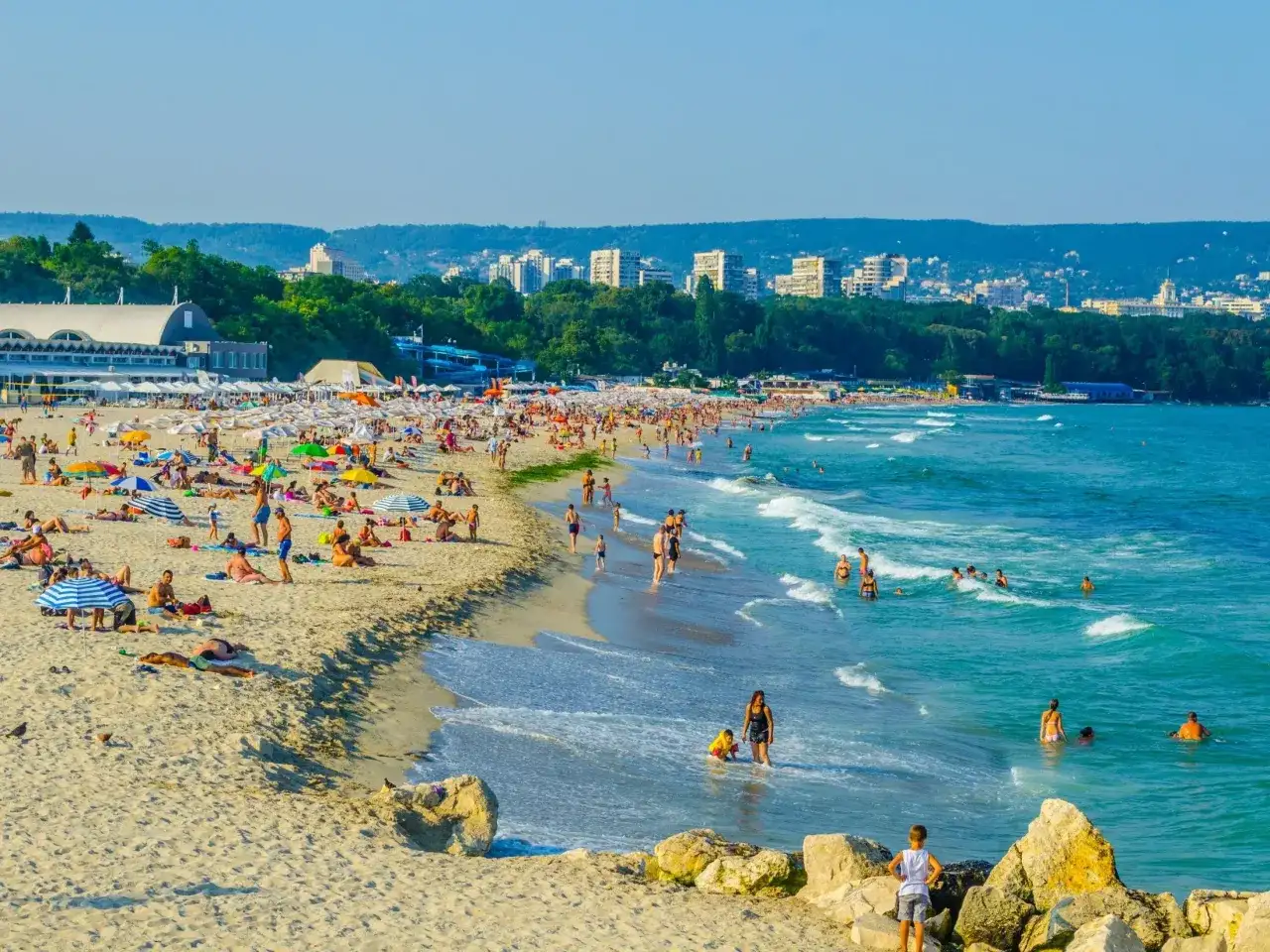Słoneczna plaża w Bułgarii, idealna na wakacje last minute all inclusive. Tłumy ludzi opalają się i kąpią w morzu.