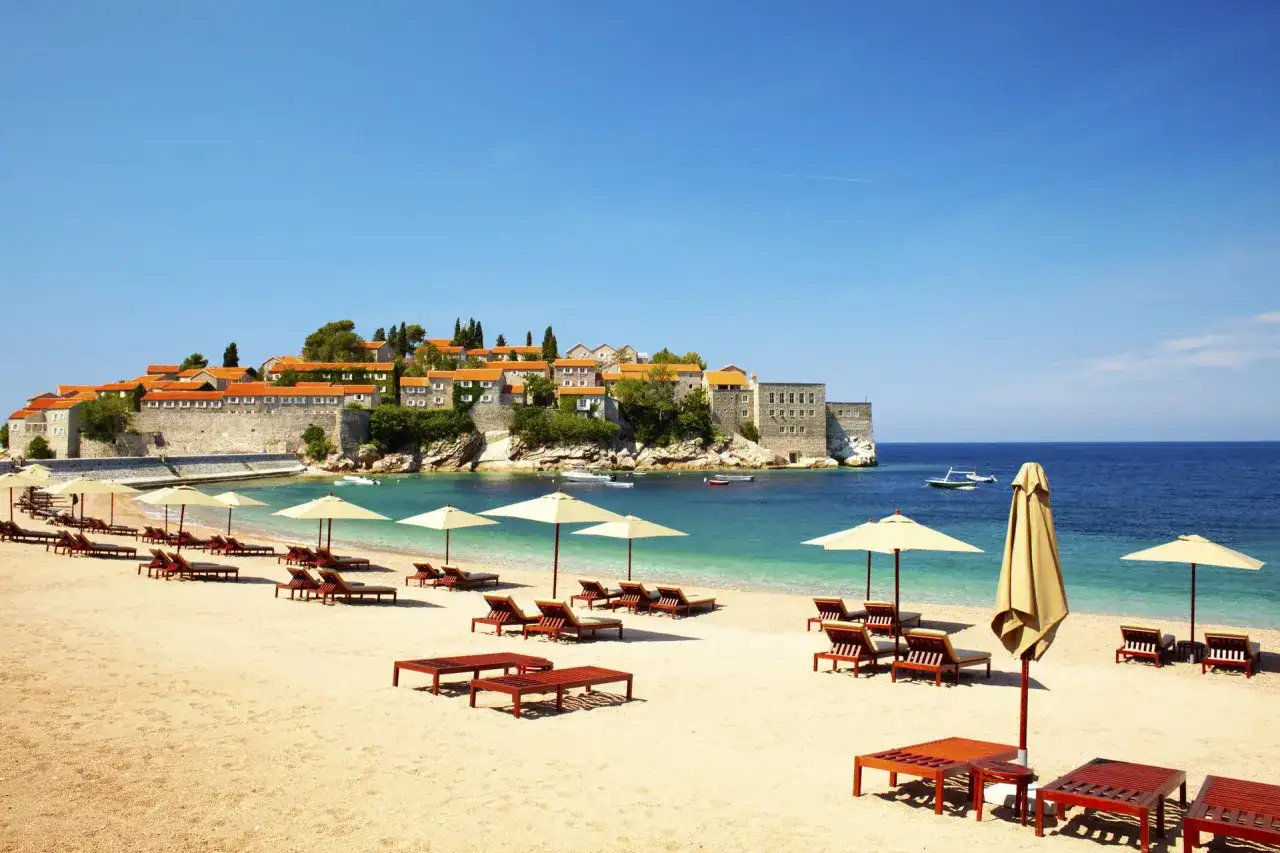 Idealne czarnogóra wakacje all inclusive: piaszczysta plaża, leżaki, parasole i widok na urokliwe miasteczko na wyspie.
