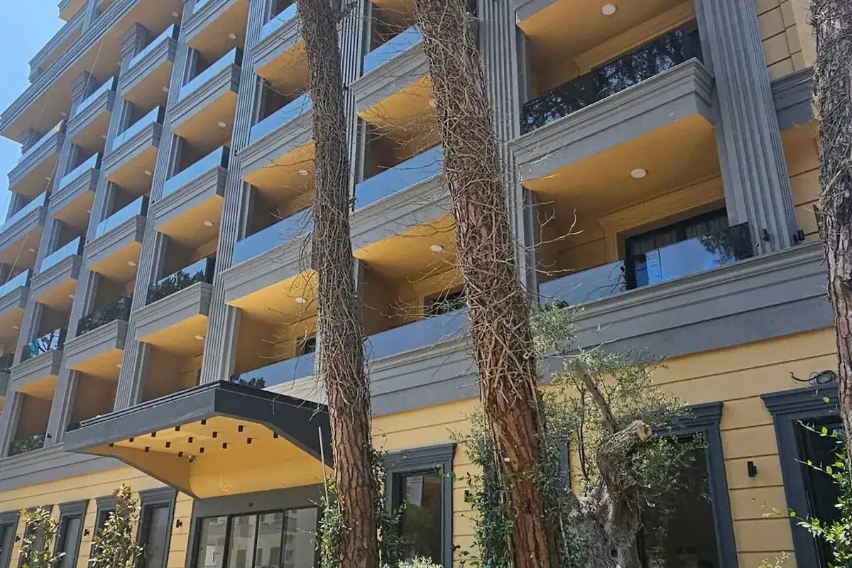 Nowoczesny hotel z żółtymi balkonami i szklanymi balustradami, otoczony drzewami. Fafa Sun Albania wita słońcem.