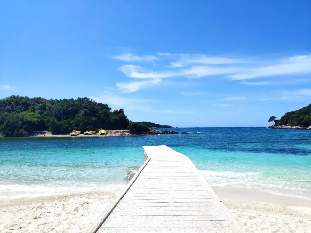 Drewniane molo prowadzi do krystalicznie czystej wody na Ksamil Beach. W oddali widać zielone wyspy i łodzie.