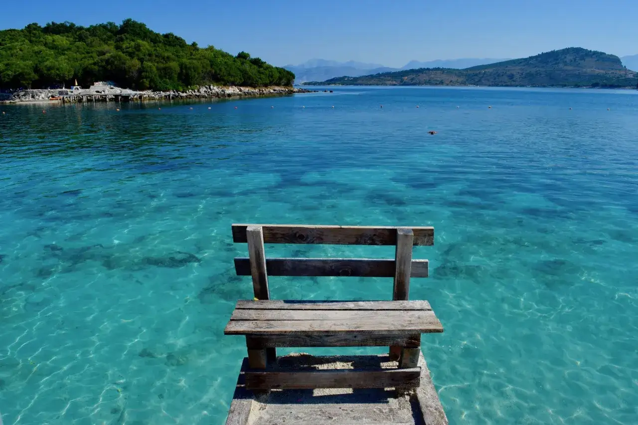 Drewniana ławka na pomoście nad krystalicznie czystą wodą. Widok na zielone wzgórza i góry w oddali. Idealne miejsce na relaks na plażach Albanii, niedaleko Tirany.