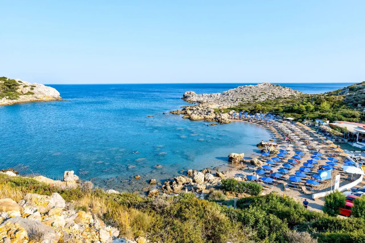Piękna zatoka z plażą i rzędami niebieskich parasoli, idealna na wakacje w Cathrin Hotel Rodos.