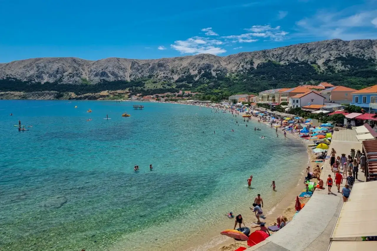 Słoneczna plaża w Baška Chorwacja. Ludzie kąpią się w turkusowej wodzie, opalają na piasku i spacerują wzdłuż wybrzeża.