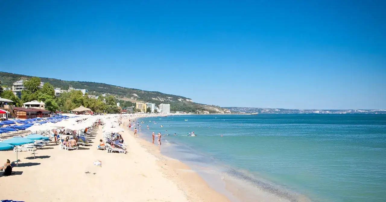 Słoneczna plaża z leżakami i parasolami, ludzie kąpią się w turkusowym morzu. W oddali widać zielone wzgórza i budynki. Kolory, co zobaczyć!