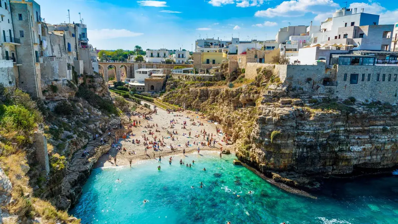 Słoneczne bari plaże z turkusową wodą, skalistymi klifami i białymi budynkami. Ludzie odpoczywają na kamienistej plaży i kąpią się w morzu.