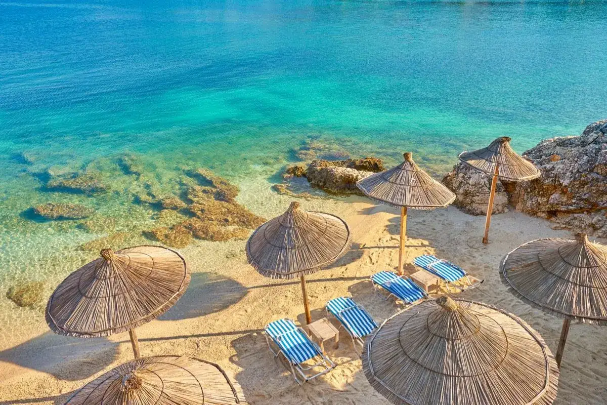 Albania - wczasy samolotem all inclusive. Słoneczna plaża z leżakami i parasolami, turkusowe morze i skały. Idealne miejsce na relaks.