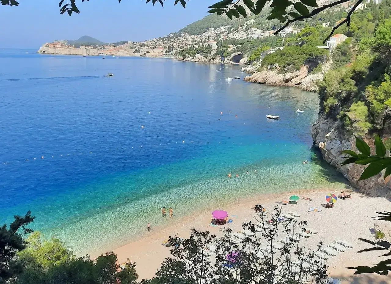 Słoneczna plaża w okolicy Dubrownika z turkusową wodą, skalistymi klifami i widokiem na zabytkowe miasto.