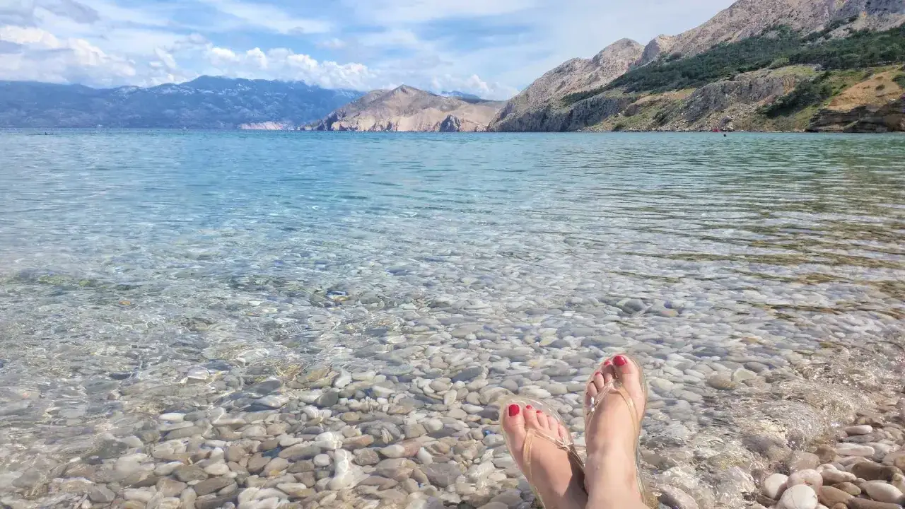 Stopy w klapkach zanurzone w krystalicznie czystej wodzie na kamienistej plaży. W tle góry i błękitne niebo. Idealny dzień na wyspie Krk.