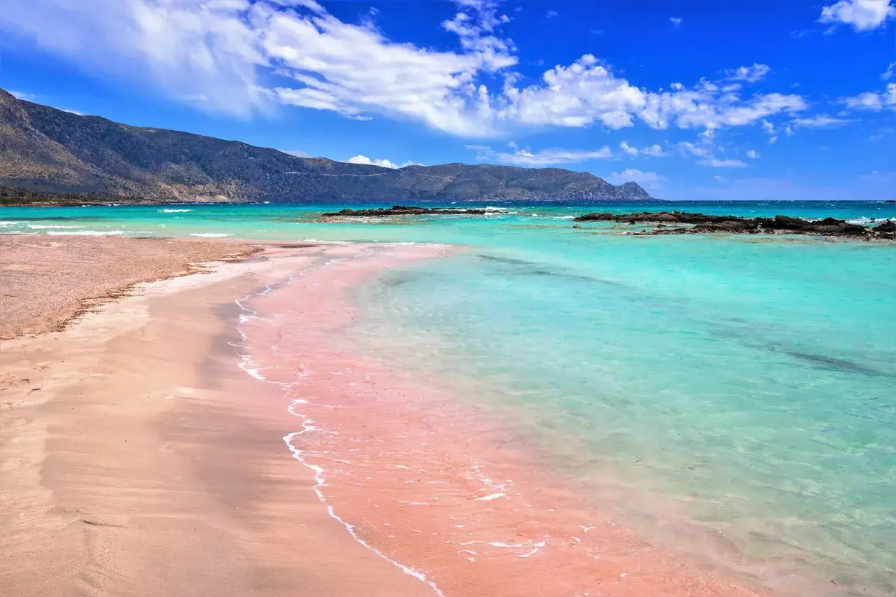 Różowa kreta plaża Elafonisi z turkusową wodą i górami w tle.