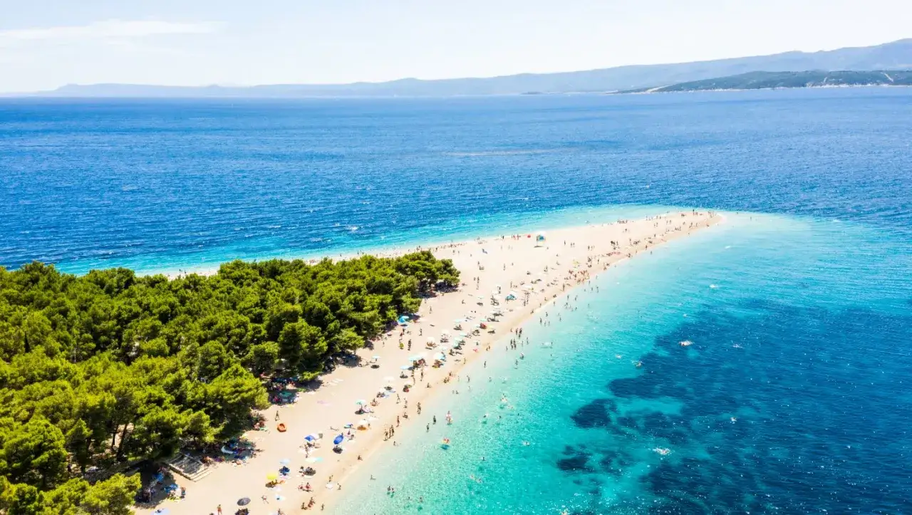 Słynna plaża w kształcie rogu obfitości, otoczona sosnami i krystalicznie czystą wodą. Wiele osób odpoczywa na piasku i pływa w morzu.