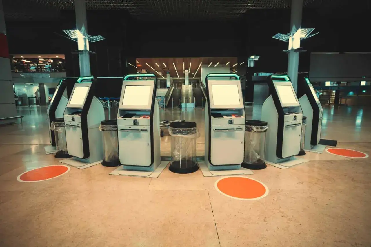 Rząd samoobsługowych stanowisk do self check-in na lotnisku. Puste ekrany czekają na pasażerów.