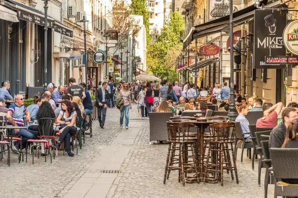 Tętniąca życiem ulica w Bukareszcie, pełna ludzi siedzących w kawiarniach i restauracjach. W tle widać zabytkowe budynki.