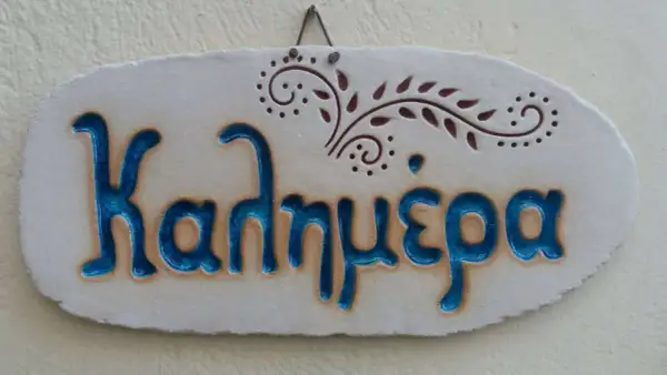 Ceramiczna tabliczka z napisem "Καλημέρα" (do zobaczenia po grecku) i ozdobnym motywem roślinnym.