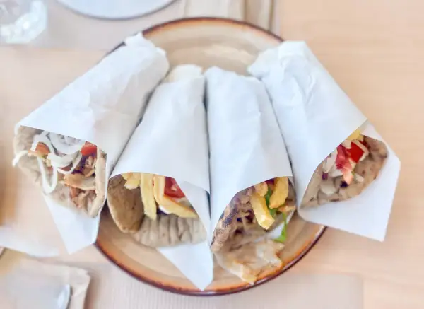 Cztery pyszne souvlaki gyros z mięsem, frytkami, warzywami i sosem, zawinięte w papier, na talerzu.