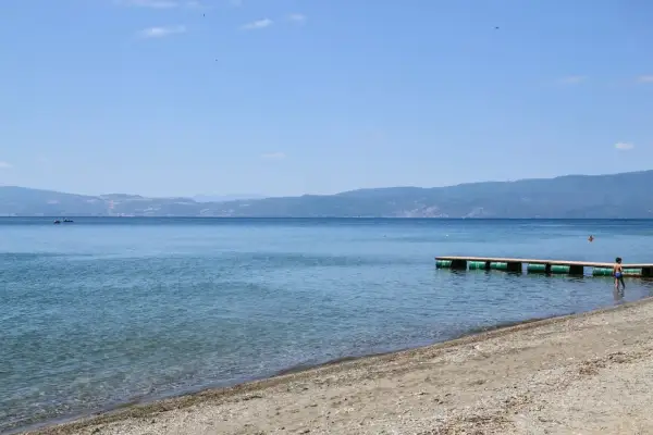 Spokojne jezioro Ochrydzkie z piaszczystą plażą i drewnianym pomostem. W oddali góry, na wodzie widać kajak.