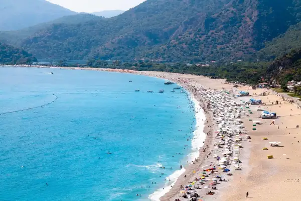 Tłum ludzi wypoczywa na piaszczystej plaży nad turkusowym morzem egejskim, otoczonej zielonymi wzgórzami.