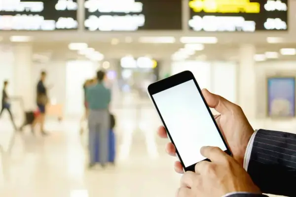 Ręka trzyma smartfon z pustym ekranem, gotowa do okazania lotniczego biletu elektronicznego. W tle lotnisko z tablicami odlotów i ludźmi.