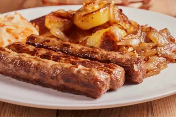 Trzy soczyste ćevapi, klasyka kuchni czarnogórskiej, podane z pieczonymi ziemniakami i cebulą oraz surówką.