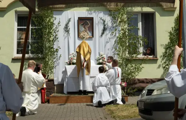 Uroczystość Bożego Ciała w długi weekend czerwiec. Procesja przed domem, kapłan w złotej szacie, wierni klęczą.