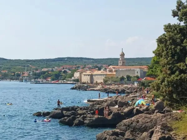 Słoneczny dzień na wyspie Krk. Ludzie odpoczywają na skalistej plaży, kąpią się w morzu. W tle widać miasteczko z charakterystyczną wieżą.