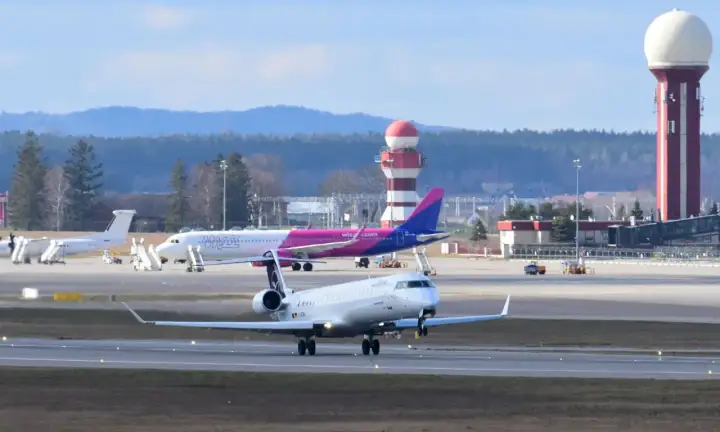 Samolot kołuje po płycie lotniska, gotowy do pierwszego lotu samolotem. W tle różowy samolot Wizz Air i wieża kontroli lotów.
